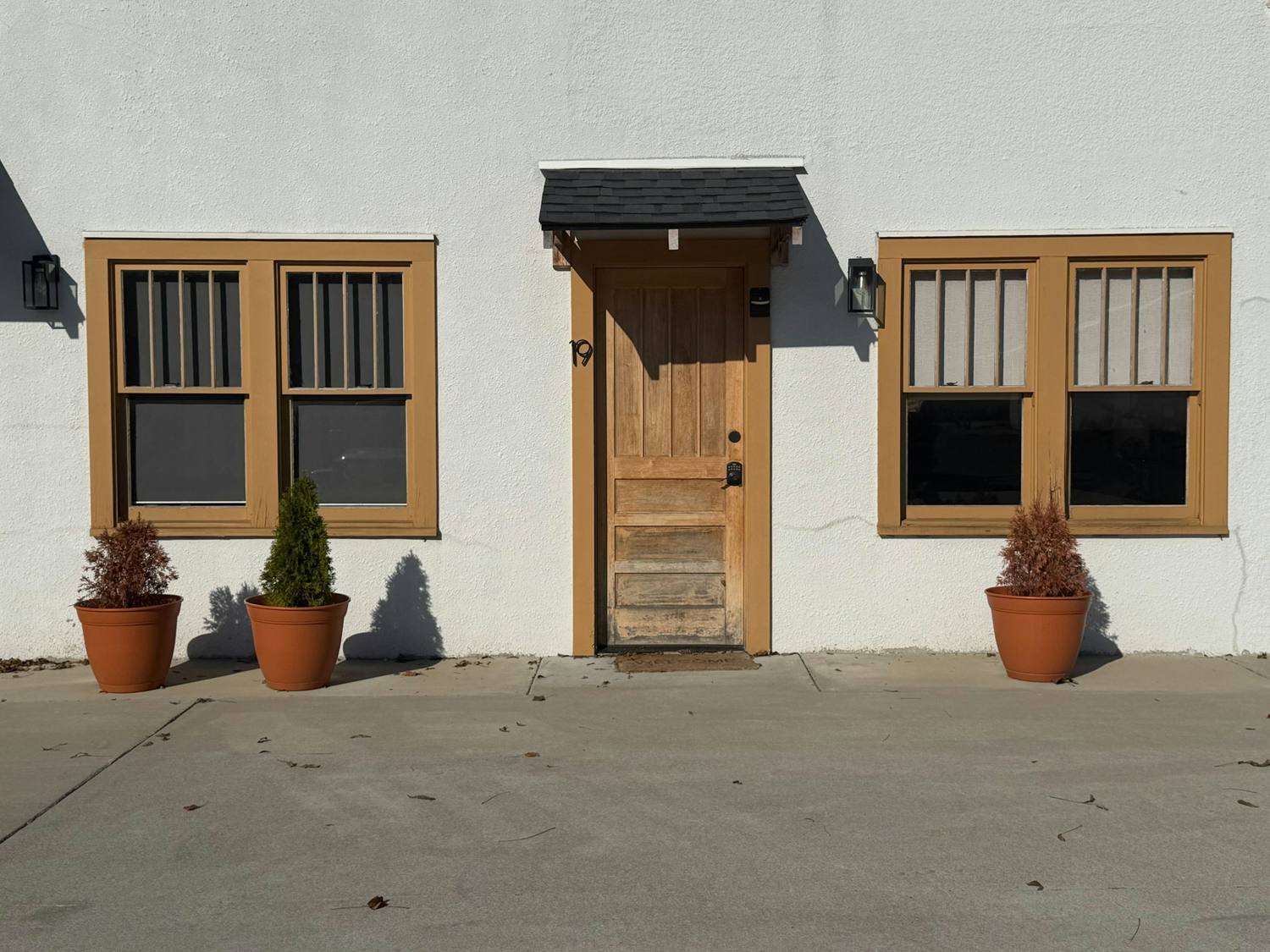 Wood entry door under a small shed roof between two windows in a white stucco wall