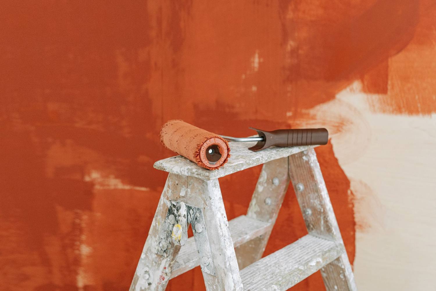 Paint roller resting on a step ladder in front of a half-painted orange wall